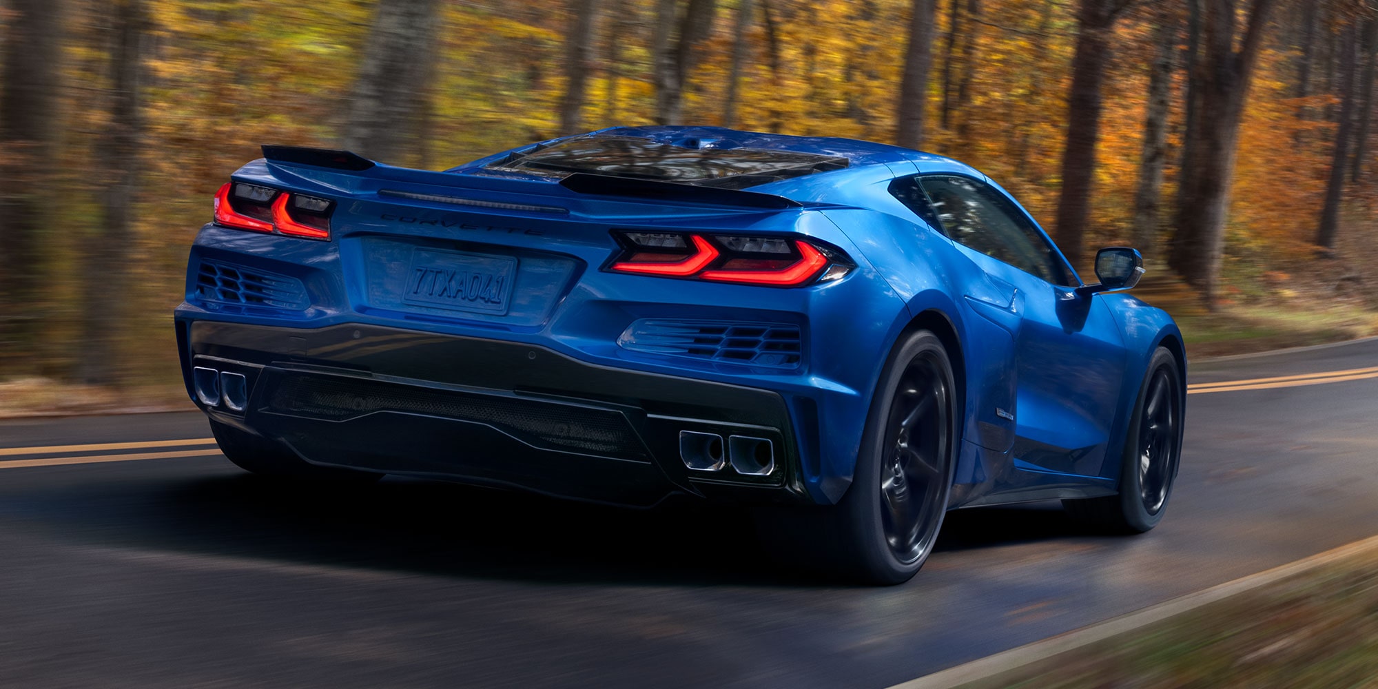 Three Quarters Rear View of a Blue Chevy Corvette E-Ray Driving Down the Road Surrounded by Trees