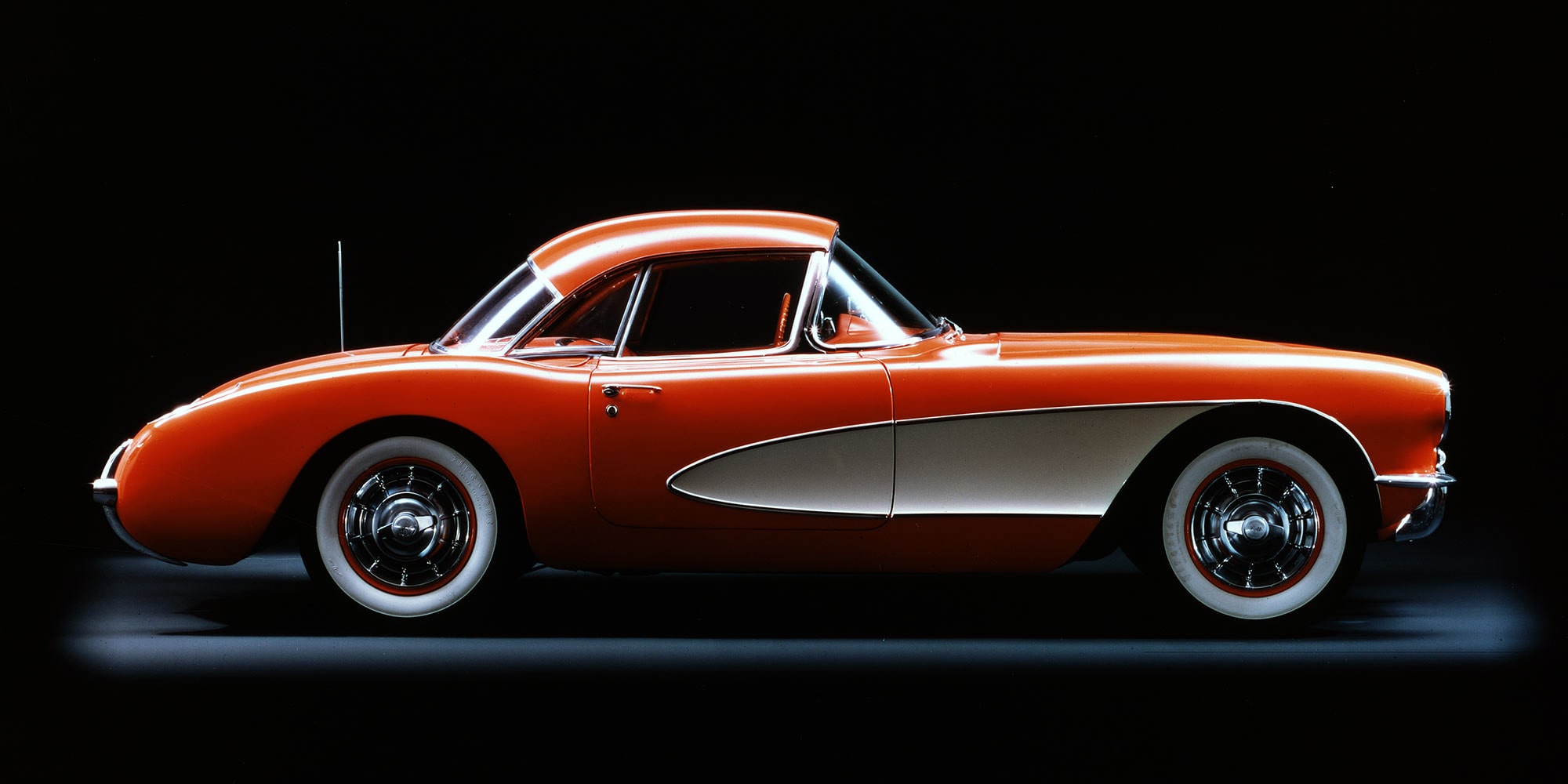 Seitenansicht eines orange-weißen Corvette C1 vor schwarzem Hintergrund.