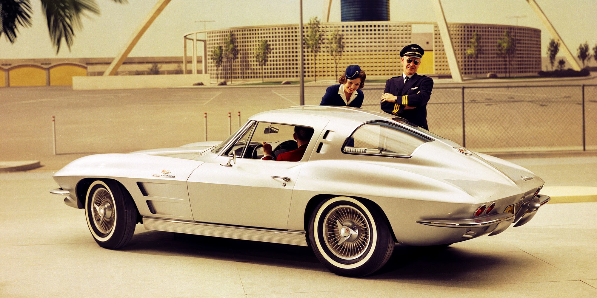 Dreiviertel-Heckansicht eines silbernen Corvette C2 Coupés, im Freien geparkt, im Gespräch mit einer Flugzeugcrew.