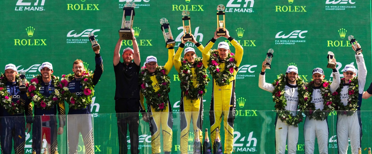 With garlands of flowers around their necks, the triumphant Corvette drivers holds their trophies overhead, against a background of sponsor’s names.